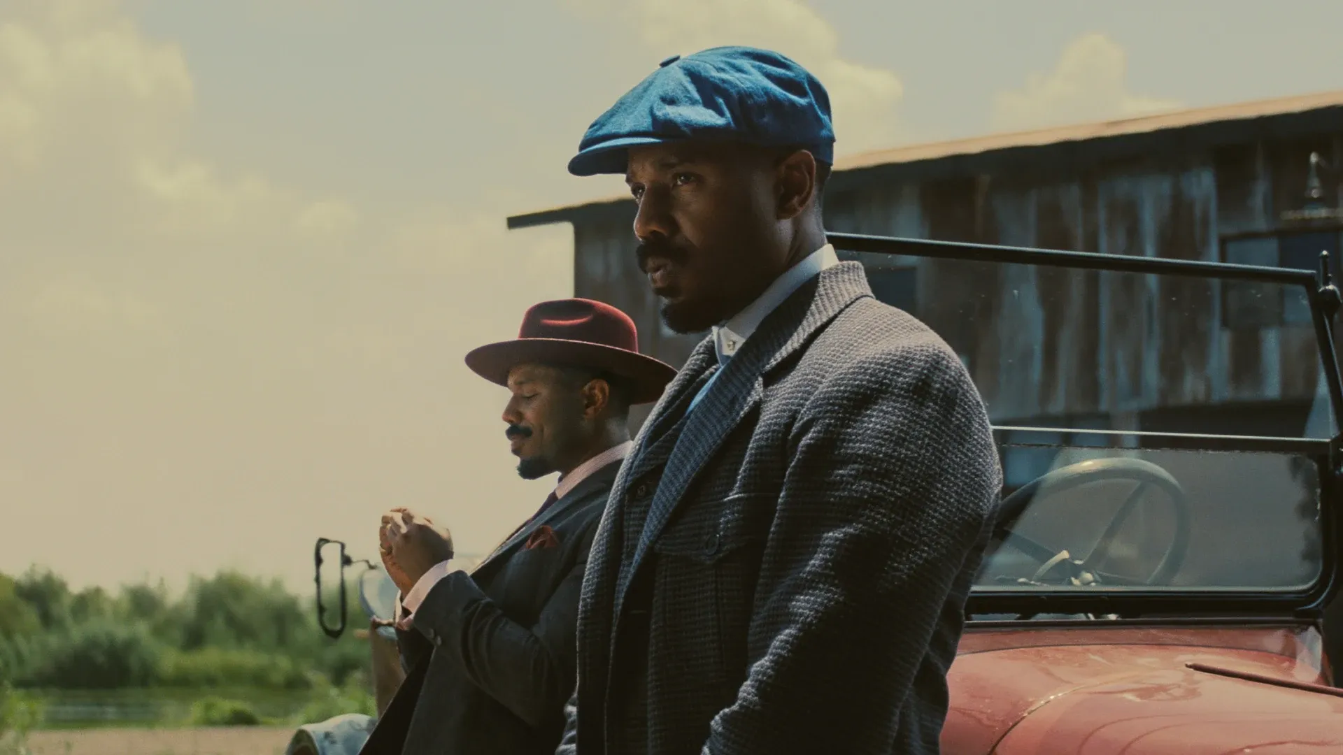 Twin brothers Smoke and Stack, played by Michael B. Jordan, are wearing hats and leaning on a vintage car in the film Sinners.